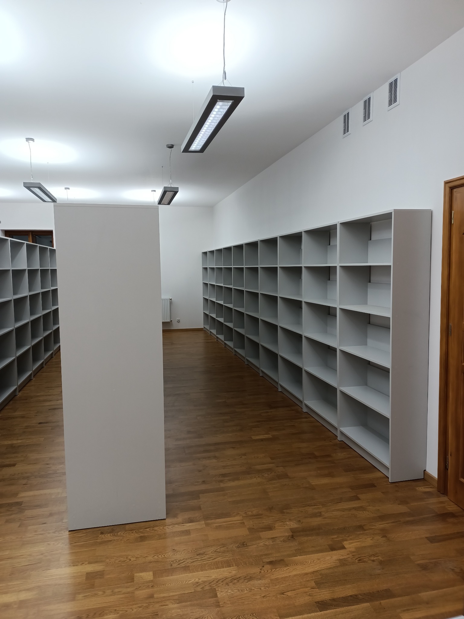Pusta biblioteka z rzędami szarych regałów na książki, oświetlona podłużnymi lampami sufitowymi, z drewnianą podłogą i białymi ścianami.