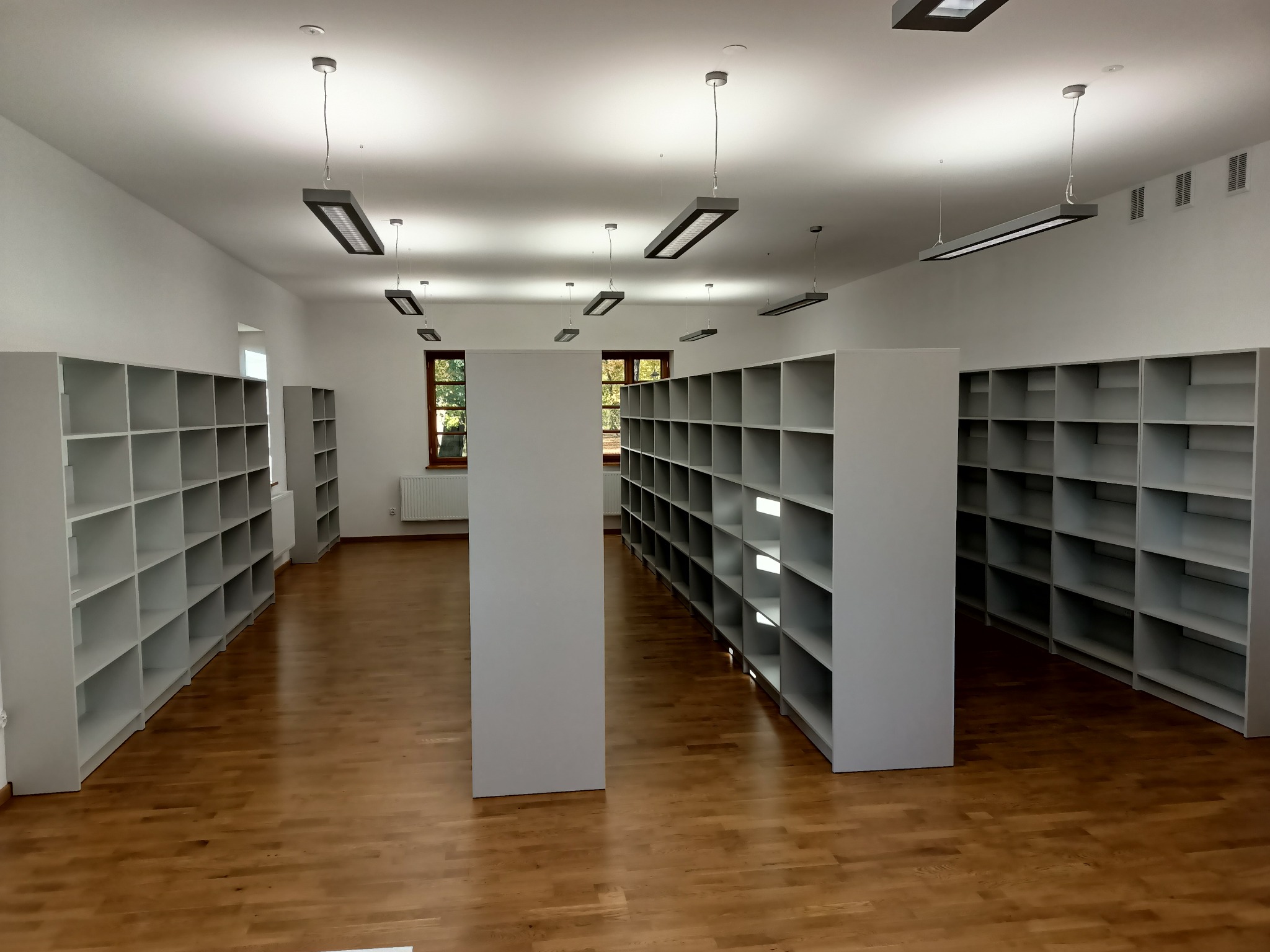 Pusta przestrzeń z rzędami białych regałów z półkami, podwieszane lampy sufitowe, parkiet na podłodze i okna z widokiem na zieleń na zewnątrz.