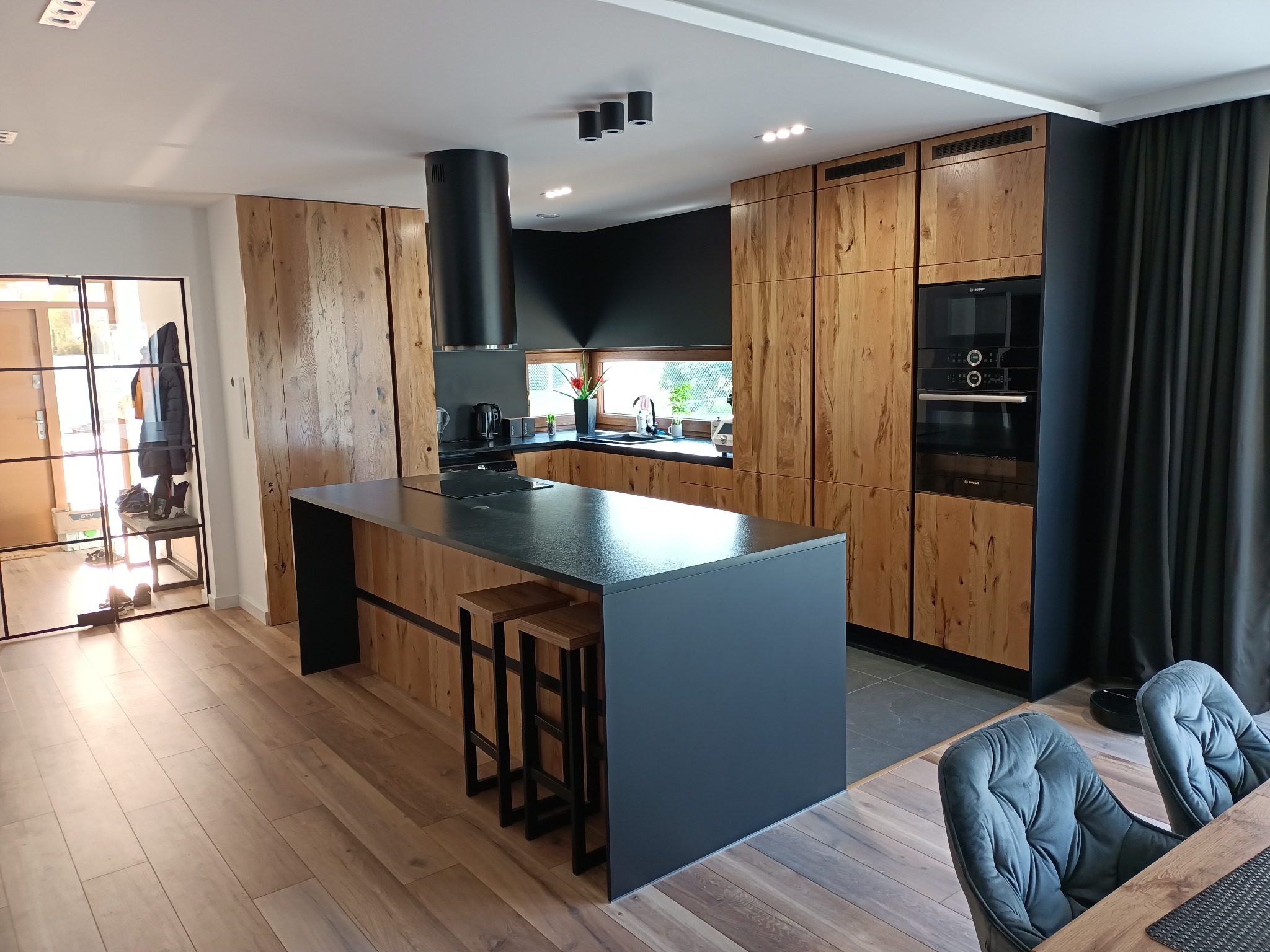 Nowoczesna kuchnia z drewnianymi szafkami, czarnym blatem wyspy i okapem, oświetlona punktowymi lampami sufitowymi, z widocznymi dwoma drewnianymi hokerami przy wyspie oraz częścią jadalni...