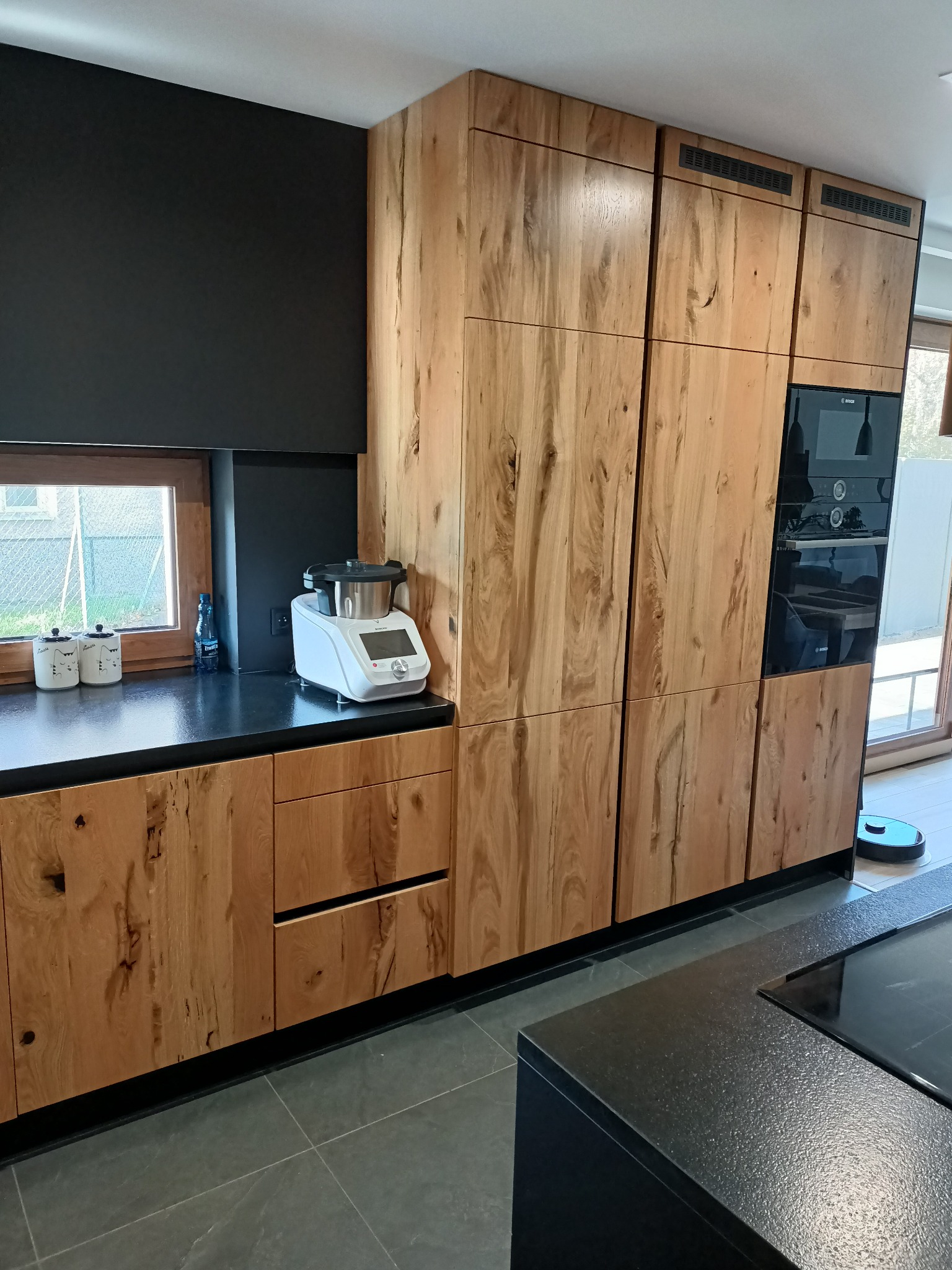 Drewniane szafki kuchenne z czarnym blatem i wbudowanymi urządzeniami w Korczynie. Widoczny robot kuchenny na blacie i robot sprzątający na podłodze.