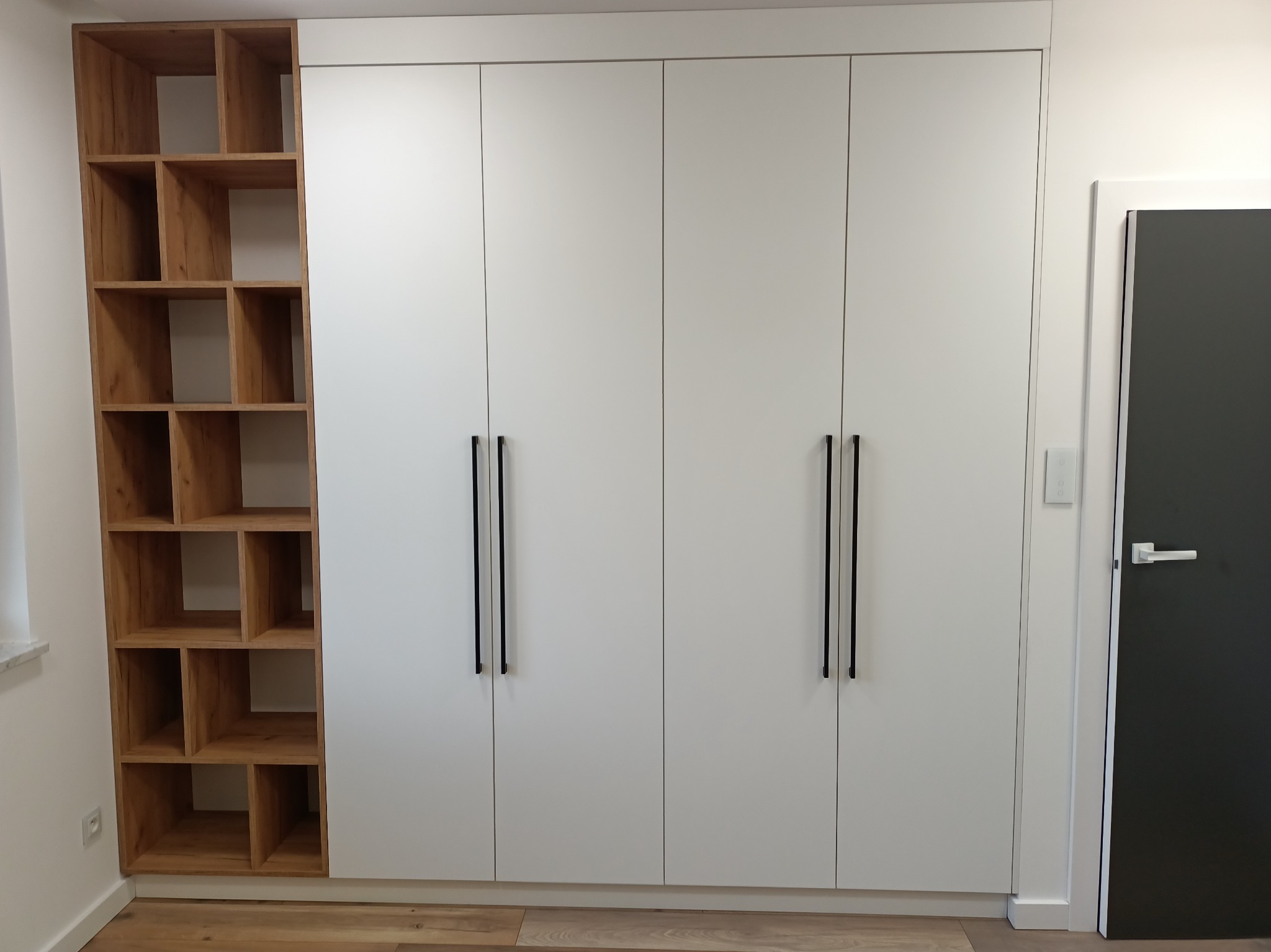Biała szafa wnękowa z czterema drzwiami i drewnianym regałem z otwartymi półkami po lewej stronie, czarne, minimalistyczne uchwyty, podłoga z paneli.
