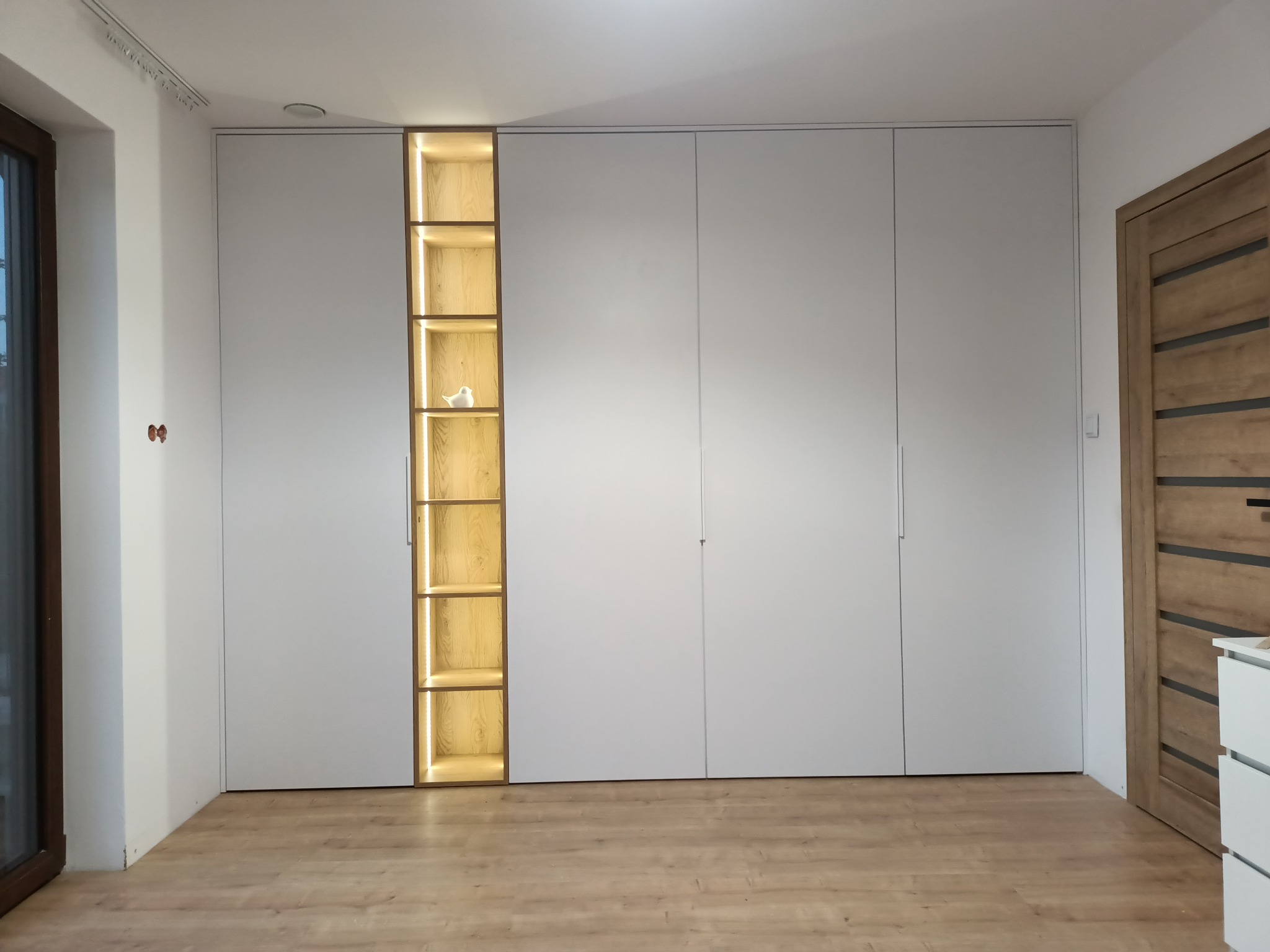 Nowoczesna szafa na wymiar z białymi frontami, wbudowana w ścianę, z podświetlaną wnęką z drewnianymi półkami, na jednej z nich stoi biała figurka ptaka.