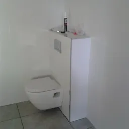 Wisząca toaleta z zabudowanym stelażem podtynkowym, białe płytki na ścianach, szare płytki podłogowe, widoczna tuba silikonu i folia na stelażu.