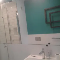 Nowoczesna łazienka z umywalką, baterią, akcesoriami i kabiną prysznicową z białymi płytkami w tle, odbijająca się w dużym lustrze z dekoracyjnym elementem na ścianie.