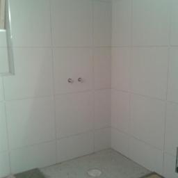 Glazurex - Wnętrze łazienki w trakcie układania białych płytek ceramicznych na ścianach, z widocznymi wyprowadzonymi rurami i poziomnicą leżącą na podłodze.