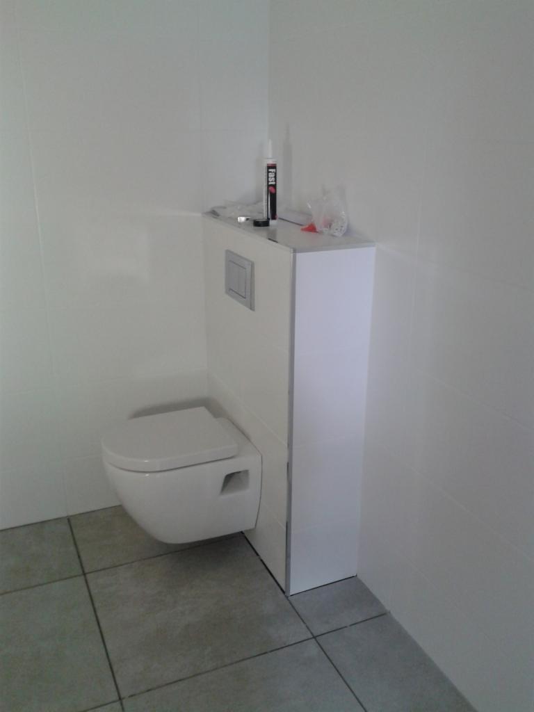 Wisząca toaleta z zabudowanym stelażem podtynkowym, białe płytki na ścianach, szare płytki podłogowe, widoczna tuba silikonu i folia na stelażu.