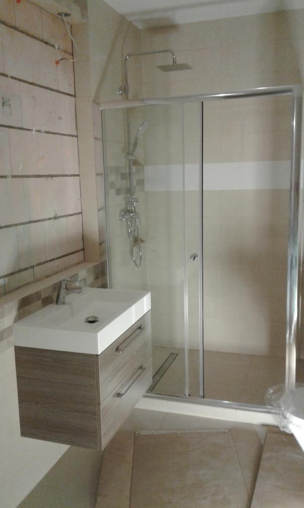 Nowoczesna łazienka w trakcie wykańczania: biała umywalka z szufladami, kabina prysznicowa z przesuwnymi drzwiami, częściowo położone płytki na ścianach.