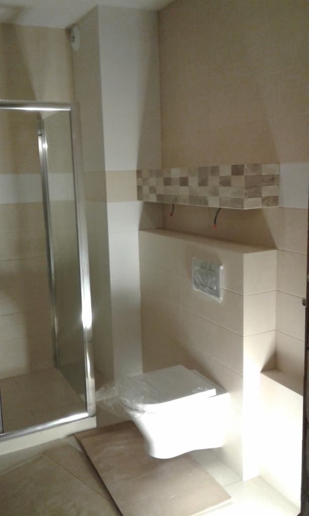 Świeżo wyremontowana łazienka z kabiną prysznicową i podwieszaną toaletą zabezpieczoną folią ochronną, wykończona beżowymi płytkami z dekoracyjnym pasem mozaiki.