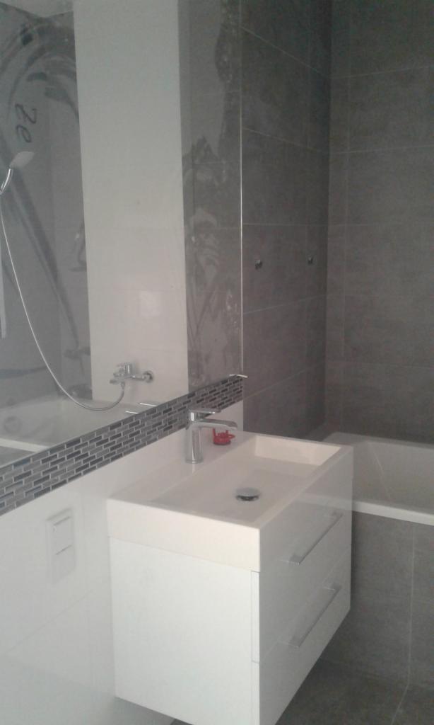 Biała umywalka z szafką z szufladami i chromowaną baterią na tle szarej ściany z płytek w nowoczesnej łazience