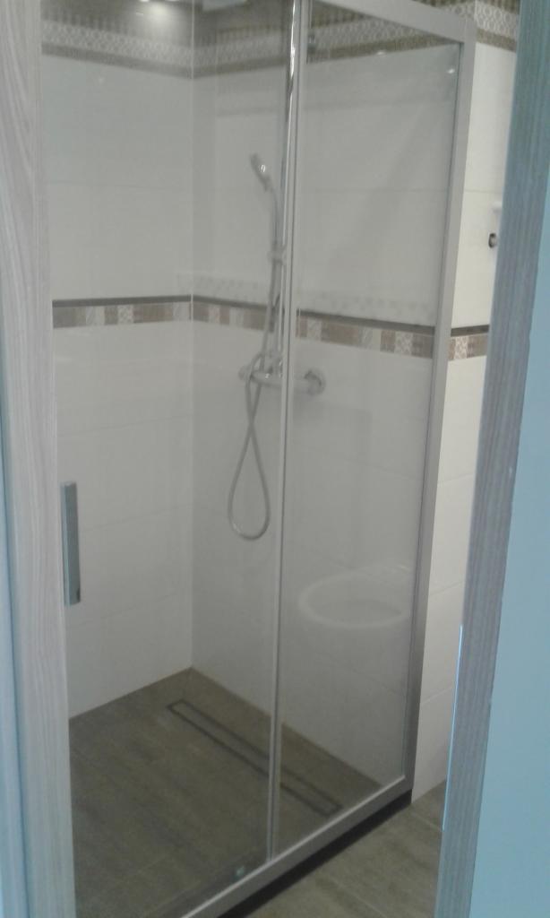 Kabina prysznicowa z przesuwnymi drzwiami, wyłożona białymi płytkami z dekoracyjnym pasem, szara podłoga z odpływem liniowym.