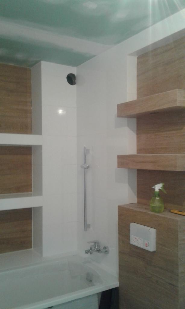 Wnętrze łazienki w trakcie wykańczania: białe płytki na ścianach, drewnopodobne panele na wnękach i półkach, sufit z płyt kartonowo-gipsowych przed malowaniem, bateria wannowa i natrysk, widoczny...