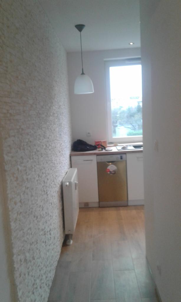 Wnętrze mieszkania po remoncie: ściana z dekoracyjną fakturą, biały grzejnik, podłoga wyłożona panelami imitującymi drewno, widok na kuchnię z białymi szafkami i złotą zmywarką, wisząca lampa...