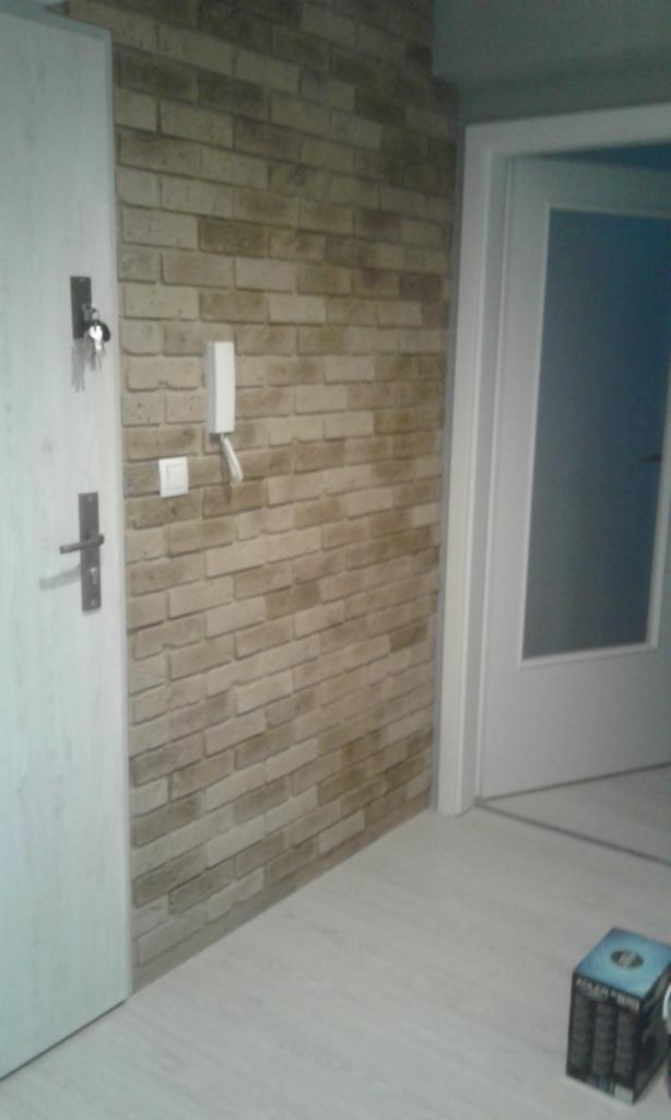 Wykończona ściana w imitacji cegły, widoczny domofon, włącznik światła, drzwi wejściowe z kluczami w zamku i drzwi wewnętrzne z szybą, podłoga z jasnych paneli.