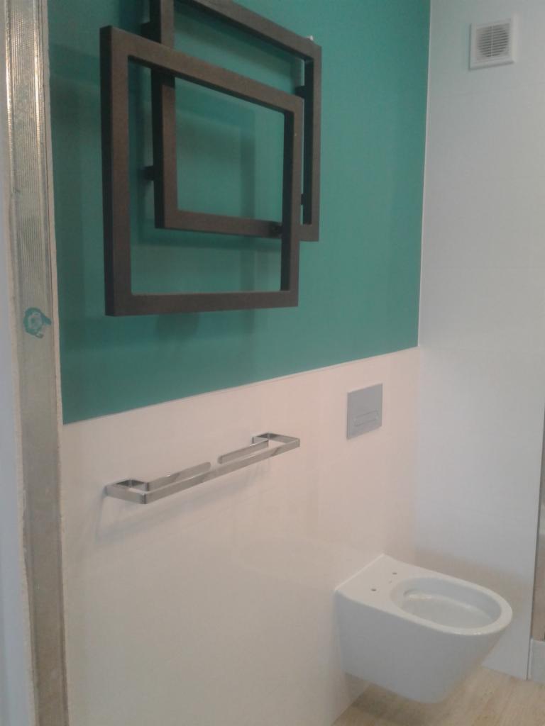 Nowoczesna toaleta podwieszana z chromowanym wieszakiem na ręczniki i dekoracyjnym grzejnikiem drabinkowym w kształcie nakładających się kwadratów na ścianie w odcieniach bieli i turkusu.