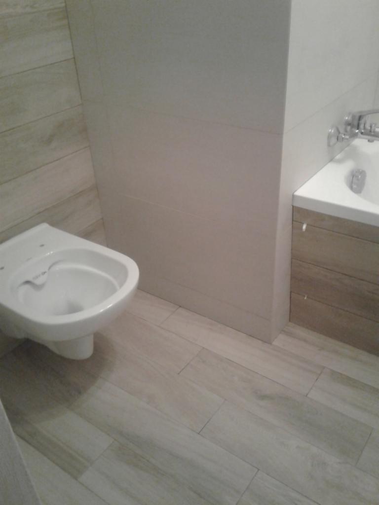 Wisząca miska WC zamontowana w łazience z płytkami imitującymi drewno na podłodze i ścianach, obok umywalka z szafką.