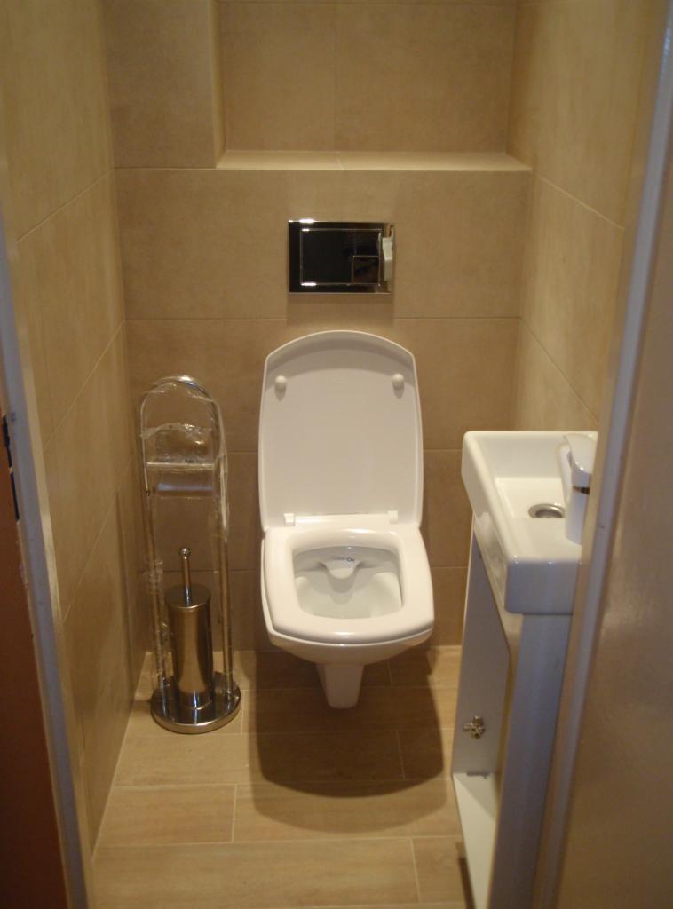 Mała toaleta z beżowymi płytkami, wiszącą muszlą klozetową, umywalką na postumencie i metalowym stojakiem na szczotkę toaletową.