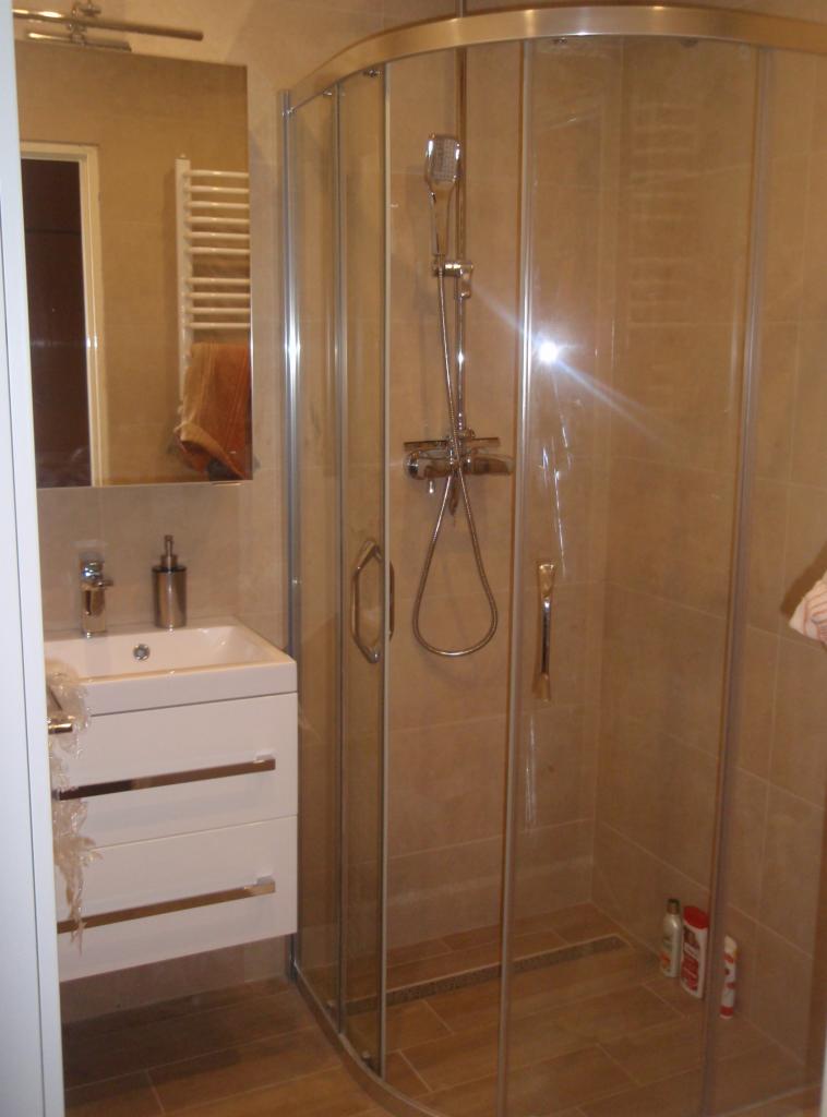 Nowoczesna kabina prysznicowa z zaokrąglonymi drzwiami, umywalka z szafką w jasnych kolorach, beżowe płytki ceramiczne na ścianach i podłodze. W tle widoczne lustro i grzejnik drabinkowy.