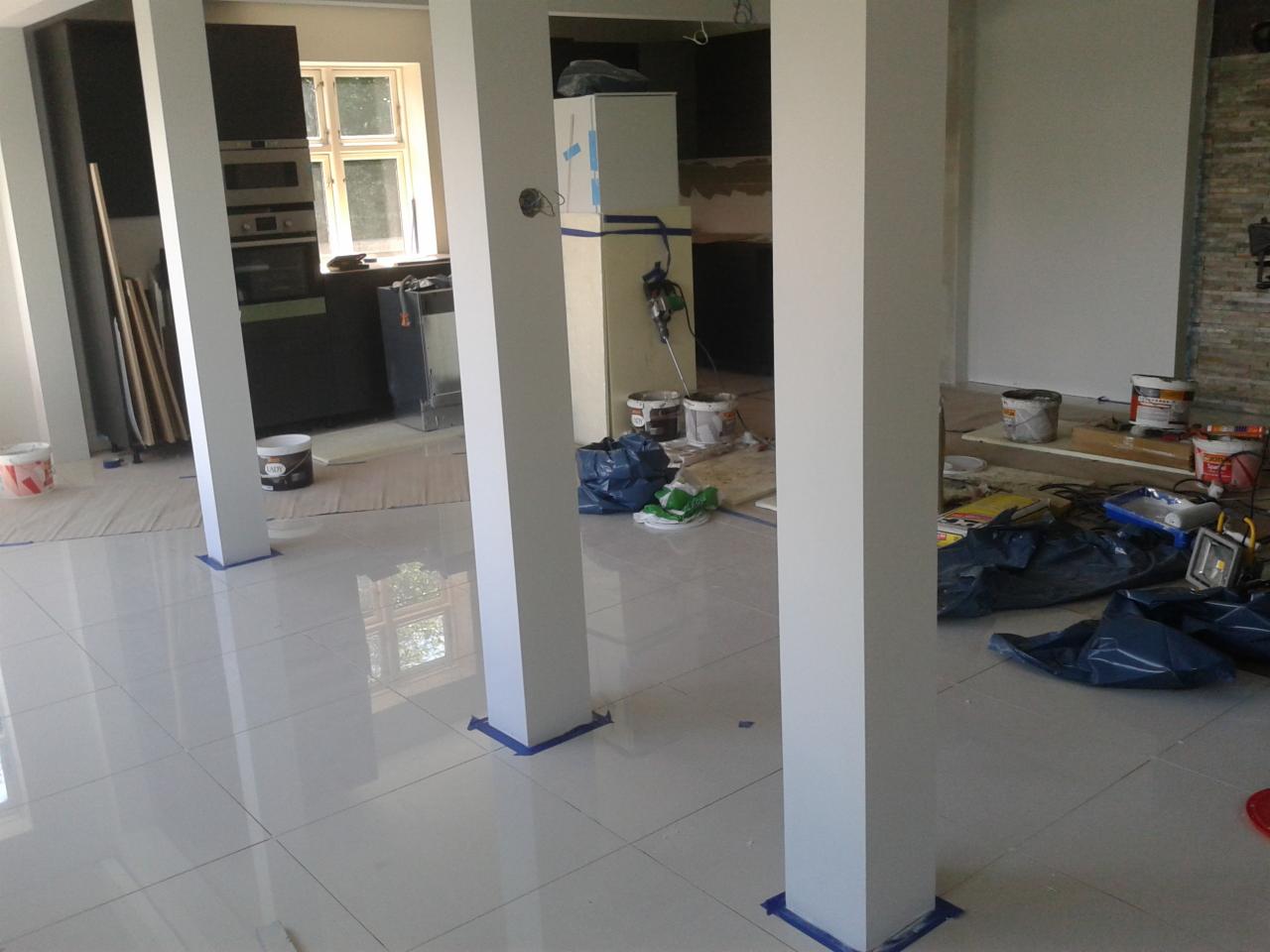 Wnętrze w trakcie prac wykończeniowych: białe, kwadratowe kolumny zabezpieczone niebieską taśmą malarską, błyszcząca podłoga z płytek, widoczne farby, narzędzia i worki na gruz, w tle kuchnia z oknem.
