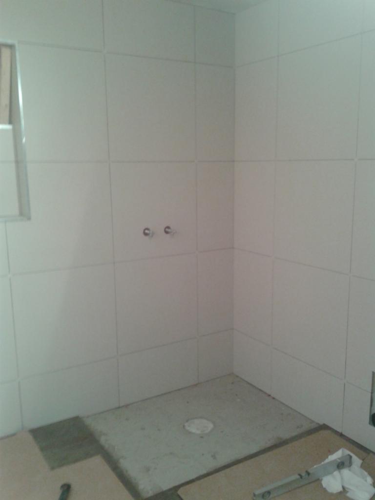 Wnętrze łazienki w trakcie układania białych płytek ceramicznych na ścianach, z widocznymi wyprowadzonymi rurami i poziomnicą leżącą na podłodze.