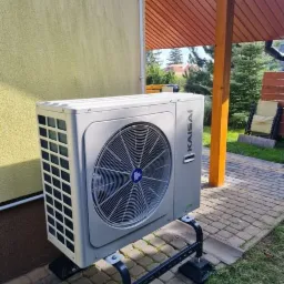 Zewnętrzna jednostka szarej pompy ciepła marki Kaisai, zamontowana na czarnej, metalowej podstawie, na tle elewacji domu, kostki brukowej i zieleni ogrodu.