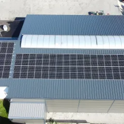 Widok z góry na zainstalowane panele słoneczne na dachu hali w Busko-Zdroju, z widocznym świetlikiem tunelowym i niebieskimi kontenerami w tle.