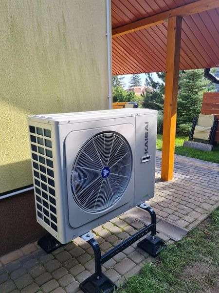Zewnętrzna jednostka szarej pompy ciepła marki Kaisai, zamontowana na czarnej, metalowej podstawie, na tle elewacji domu, kostki brukowej i zieleni ogrodu.
