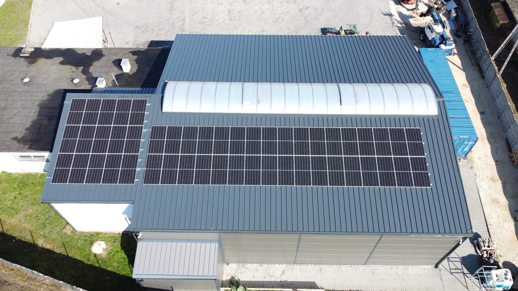 Widok z góry na zainstalowane panele słoneczne na dachu hali w Busko-Zdroju, z widocznym świetlikiem tunelowym i niebieskimi kontenerami w tle.