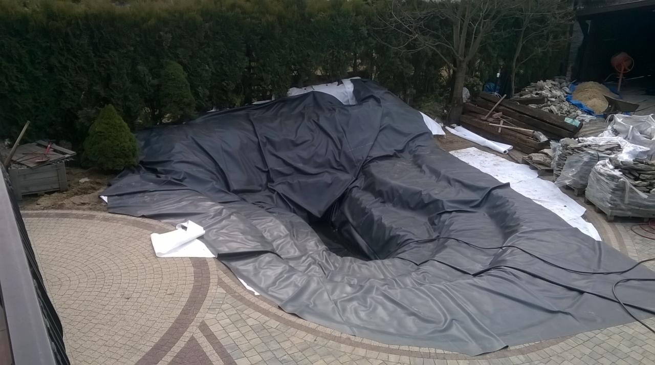 Wykopany dół pod oczko wodne wyłożony czarną folią, obok materiały budowlane, kamień i narzędzia.