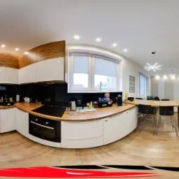 Wnętrze nowoczesnej kuchni z drewnianymi i białymi szafkami, blatem z drewna, czarnym panelem nad blatem, oknem z roletami, stołem z krzesłami barowymi i widokiem na salon