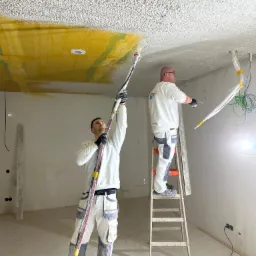 Dwóch pracowników w białych uniformach aplikuje tynk na sufit za pomocą agregatu, jeden stoi na drabinie, widoczne kable elektryczne zabezpieczone folią.