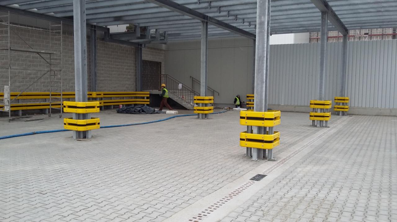 Wnętrze hali w trakcie budowy z zamontowanymi odbojnicami w kolorze żółto-czarnym na stalowych słupach, widoczni pracownicy w odblaskowych kamizelkach i rusztowania.