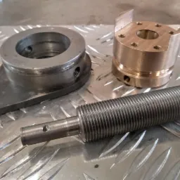 Metalowe elementy obrabiane: pierścień z podstawą, tuleja z wycięciami i gwintowany trzpień, ułożone na ryflowanej blasze.