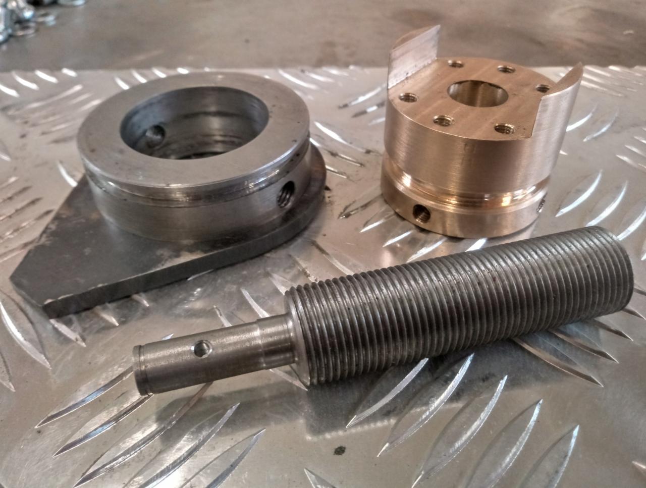 Metalowe elementy obrabiane: pierścień z podstawą, tuleja z wycięciami i gwintowany trzpień, ułożone na ryflowanej blasze.