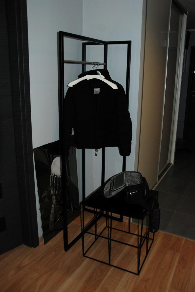 Czarny metalowy wieszak na ubrania z czarną kurtką na białym wieszaku, obok stoi metalowy stolik z torbą, a na podłodze oparta jest grafika.