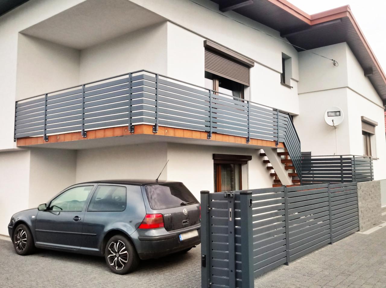 Szare, poziome metalowe ogrodzenie i balustrada balkonowa przy białym domu z szarym samochodem Volkswagen Golf zaparkowanym obok.