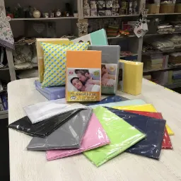 Prezentacja różnokolorowych prześcieradeł i pościeli na stole w sklepie z tekstyliami domowymi, z widocznymi opakowaniami i dekoracyjnymi elementami na półkach w tle.