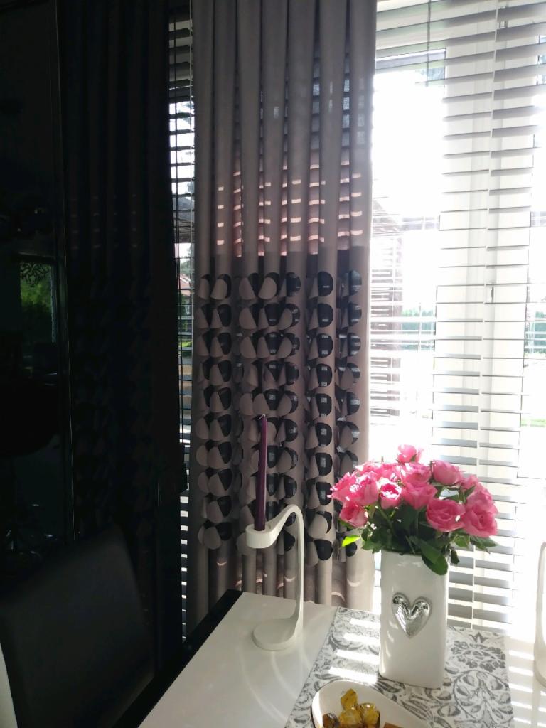 Szaro-czarne zasłony z geometrycznym wzorem w pokoju, biały stół z bukietem różowych róż w białym wazonie z srebrnym sercem, obok biały świecznik z fioletową świecą, żaluzje w oknie w tle.