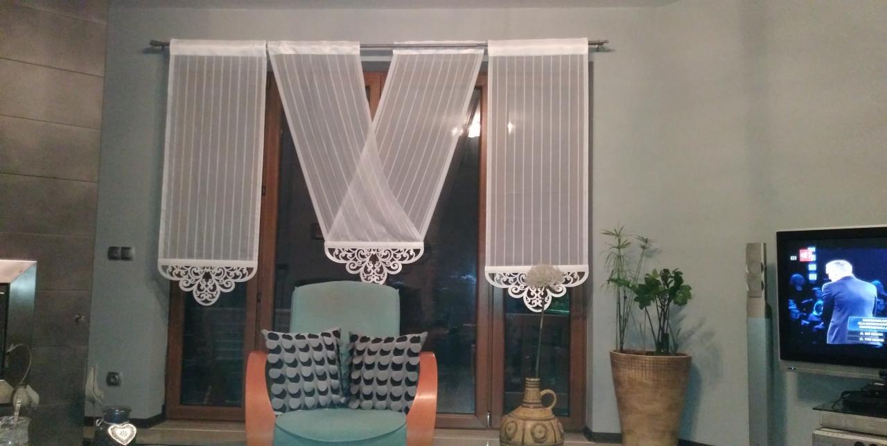 Nowoczesne, białe, transparentne panele okienne z ażurowym wykończeniem dolnej krawędzi, w salonie z turkusowym fotelem i telewizorem.