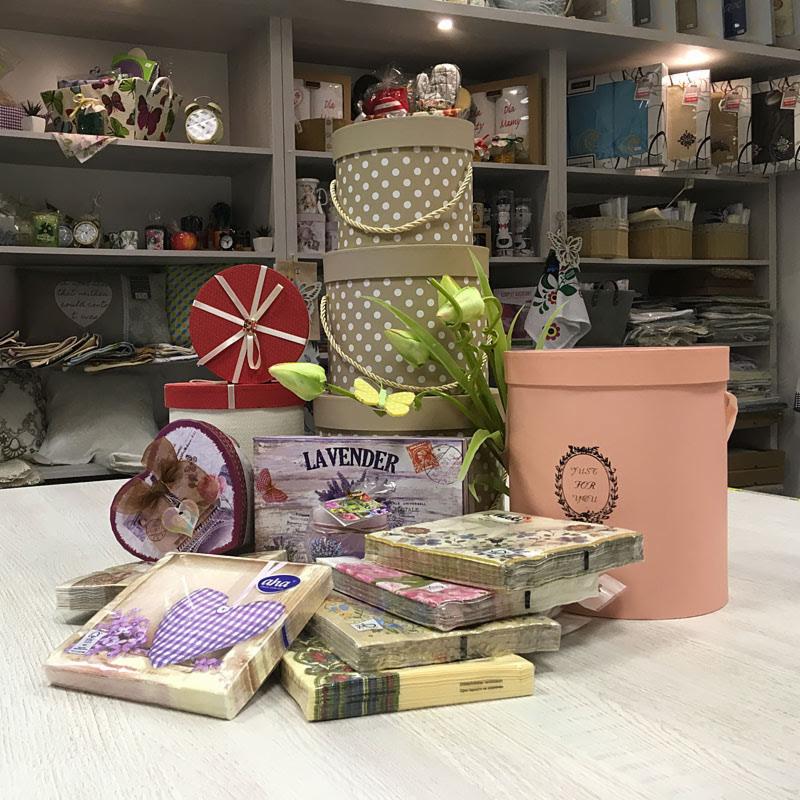 Ekspozycja różnorodnych pudełek ozdobnych, serwetek papierowych z motywami kwiatowymi i lawendowymi, oraz dekoracji na tle regałów sklepowych wypełnionych tkaninami i akcesoriami.