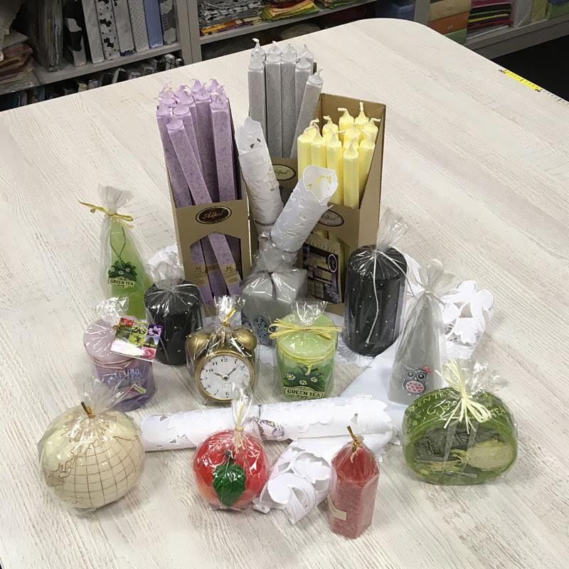 Różnorodny asortyment świec dekoracyjnych i zapachowych, w tym świece stożkowe w odcieniach fioletu, szarości i żółci, oraz świece w kształcie jabłka, kuli ziemskiej i słoika z napisem 'Green Tea',...
