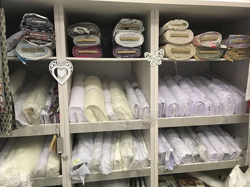 Półki w sklepie z tkaninami, wypełnione różnorodnymi rolkami materiałów, głównie białych i wzorzystych, z dekoracyjnymi elementami w kształcie serca i motyla.