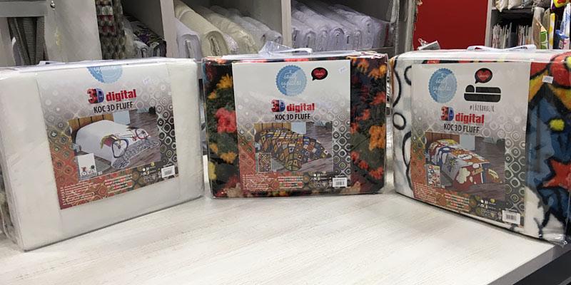 Trzy zapakowane koce 3D Fluff z różnymi wzorami (geometryczny, kwiatowy, komiksowy) stojące na jasnym blacie w sklepie z tekstyliami.