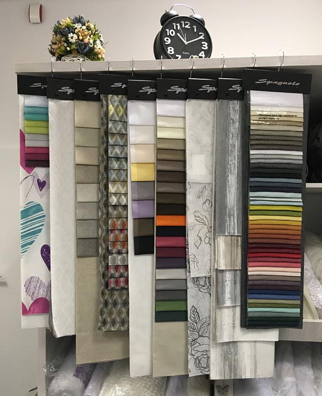 Ekspozycja próbek różnorodnych tkanin wiszących na wieszakach, z zegarem i bukietem kwiatów na górnej półce, prezentująca szeroką gamę kolorów i wzorów.