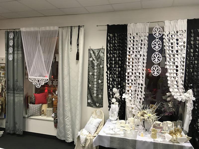 Ekspozycja różnorodnych tkanin i dekoracji okiennych w sklepie: zasłony panelowe z wycinanymi wzorami, firany woalowe z koronkowym wykończeniem, dekoracyjne poduszki na parapecie, oraz stół...