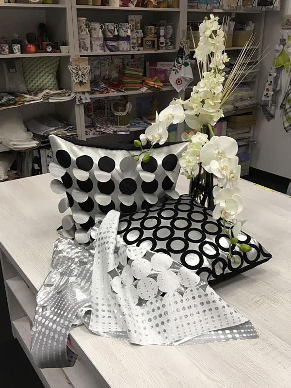 Dekoracyjne poduszki z motywem kół i srebrzysta tkanina z wypukłymi kółkami, obok wazon z białymi orchideami, na tle półki z tkaninami i akcesoriami.