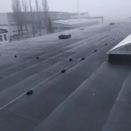 Instalacja odgromowa na płaskim dachu przemysłowym w mglisty dzień, widoczne czarne uchwyty i biały przewód, w tle budynki przemysłowe.