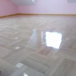 Parkiet ułożony we wzór jodełki, po nałożeniu lakieru, z widocznym odbiciem światła od okna, na tle różowej ściany poddasza.