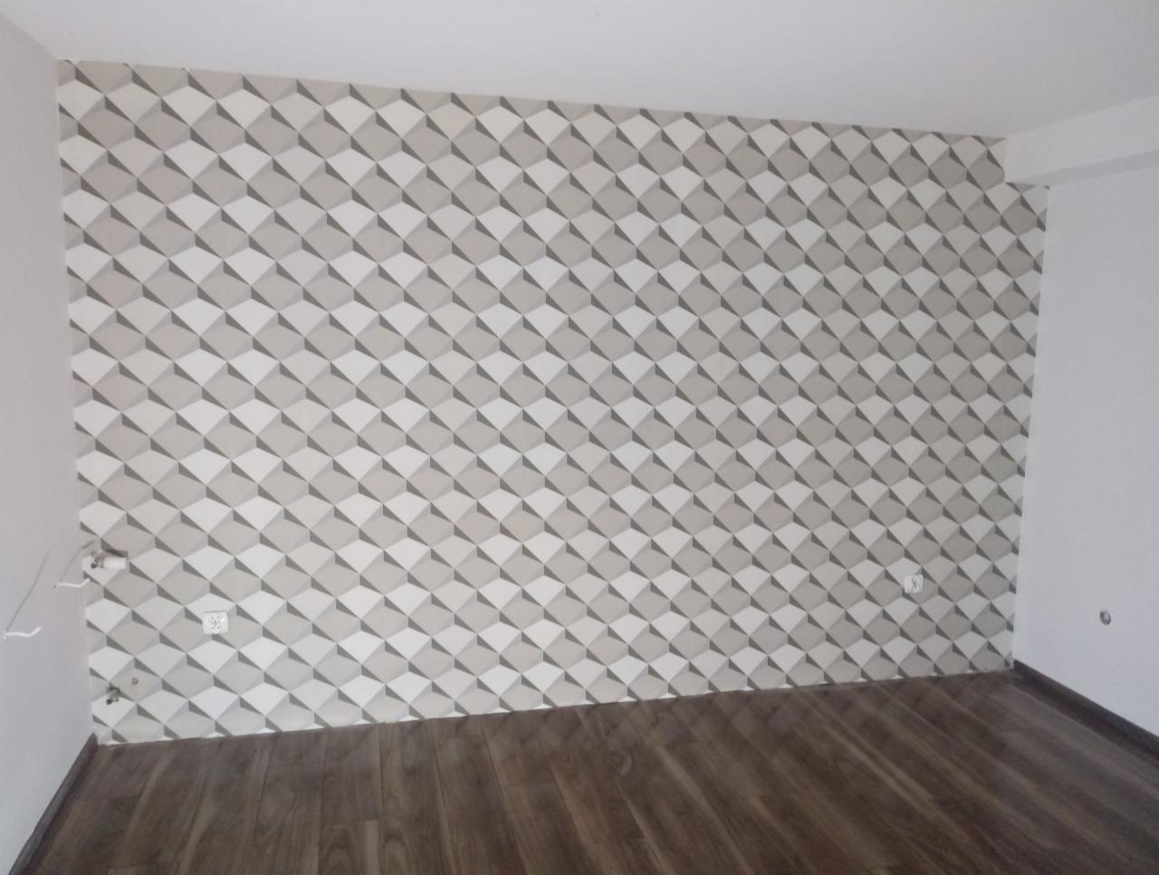Wnętrze pokoju z ułożonymi panelami podłogowymi i ścianą pokrytą tapetą w geometryczny wzór rombów w odcieniach szarości i bieli, widoczne gniazdka elektryczne na ścianie.