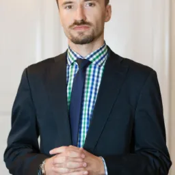 Portret adwokata w ciemnym garniturze i kraciastej koszuli, z zapiętymi dłońmi, stojącego na tle jasnych drzwi.