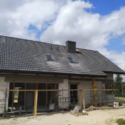 Niedokończony dom z białych bloczków, z ciemnoszarą dachówką i kominem z cegły klinkierowej, widoczne rusztowania i drewniane podpory szalunkowe, na dachu dwa okna połaciowe.